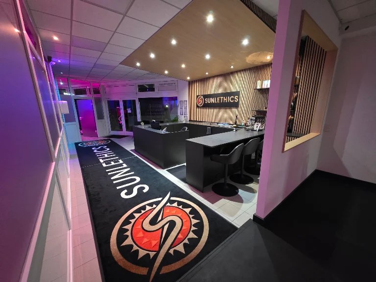 Eingangsbereich bei SUNLETHICS mit Logo-Teppich, Theke und farbiger Beleuchtung